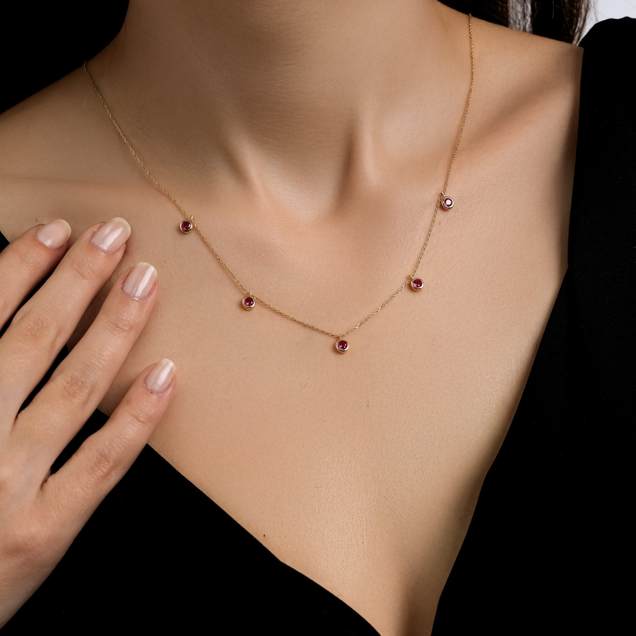 14K solid yellow gold station ruby necklace worn elegantly with a hand gently touching the neckline, showcasing five ruby stones.