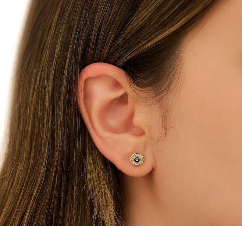 14K yellow gold minimalist evil eye stud earrings displayed on a model's ear, featuring CZ detail.