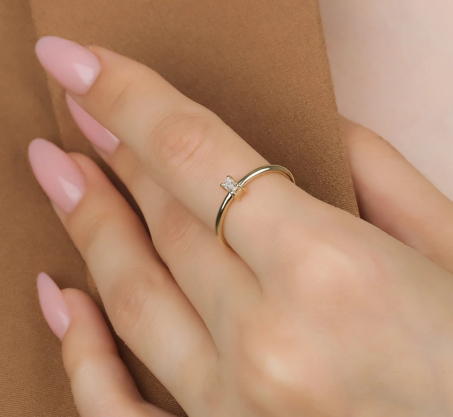 14K gold baguette diamond solitaire ring worn on a hand with light pink nails against a brown background.