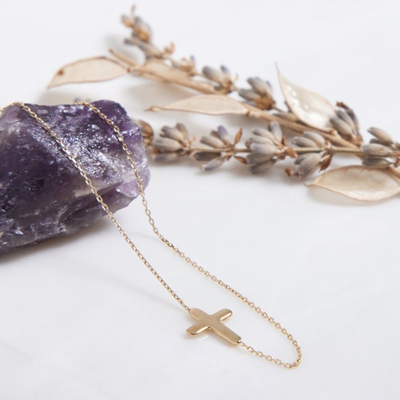 14K solid yellow gold minimalist sideways cross necklace on a white surface with lavender leaves.