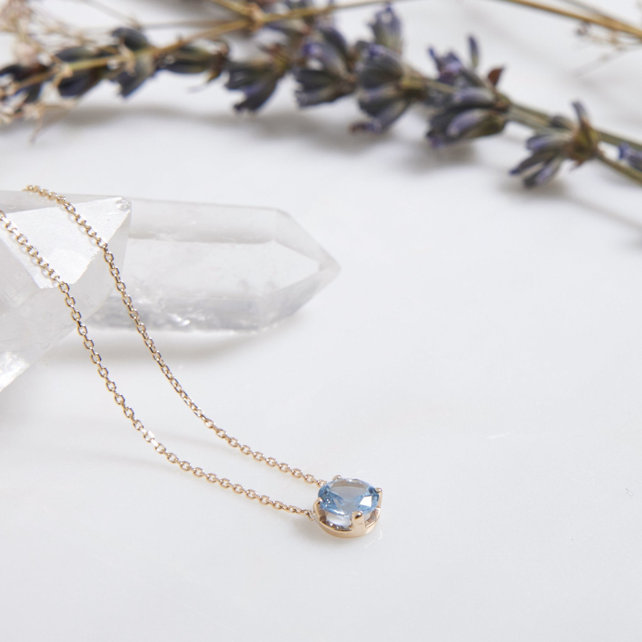 14K white gold 6mm prong setting solitaire aquamarine necklace on a crystal surface with lavender accents.