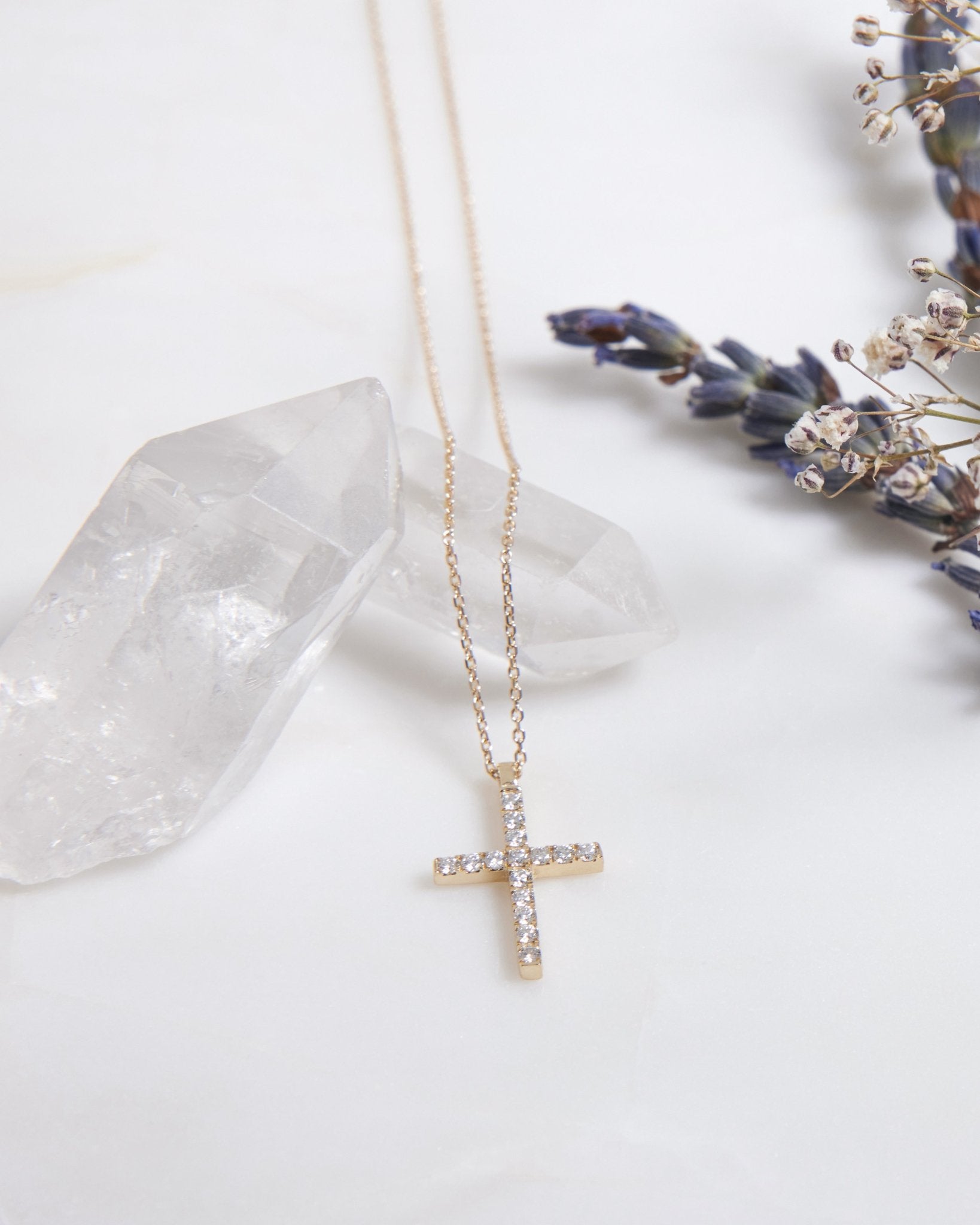14K solid white gold diamond cross necklace on a crystal surface with lavender flowers in background.