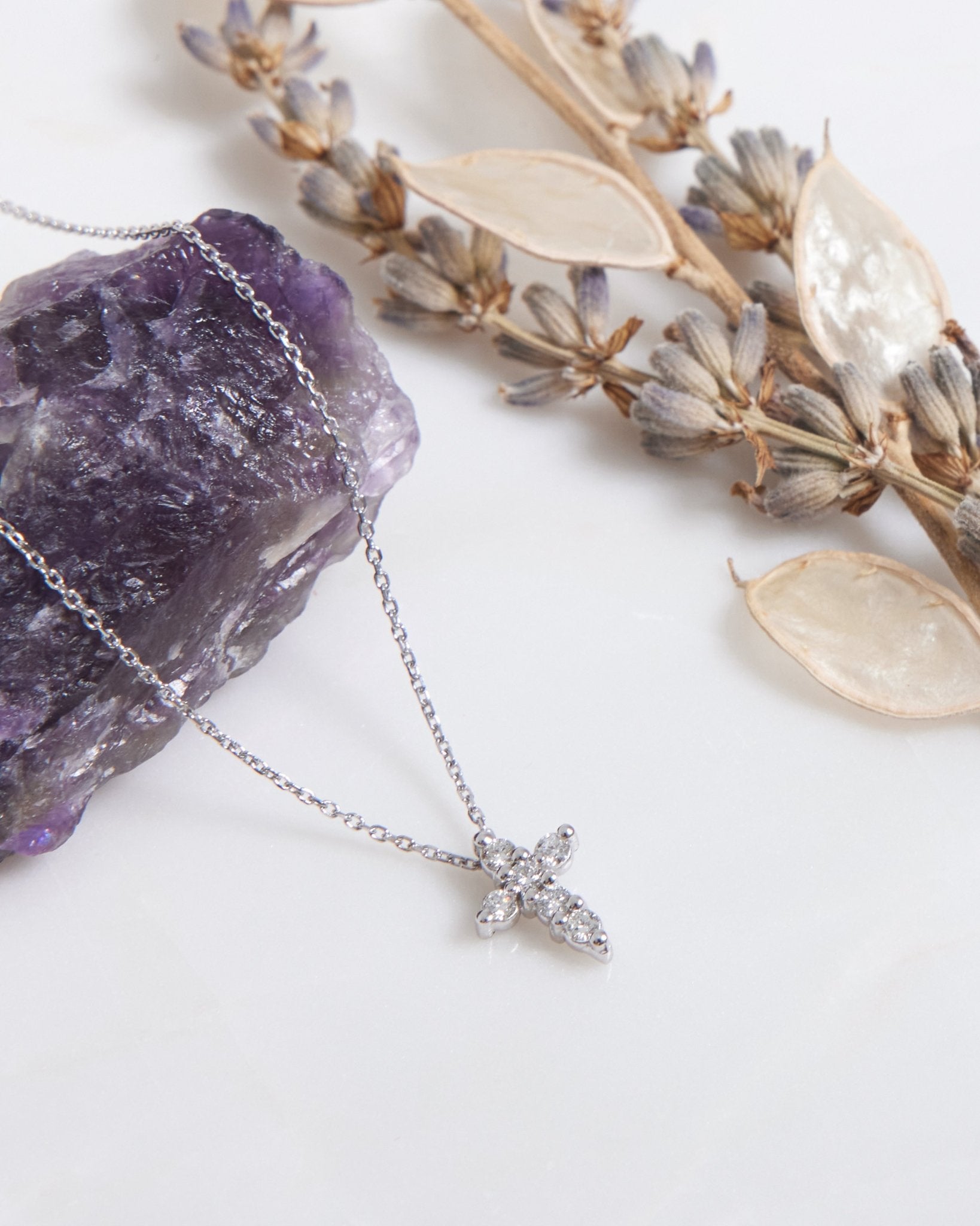 14K white gold minimalist diamond cross necklace on a white surface with purple amethyst crystal and dried flowers.