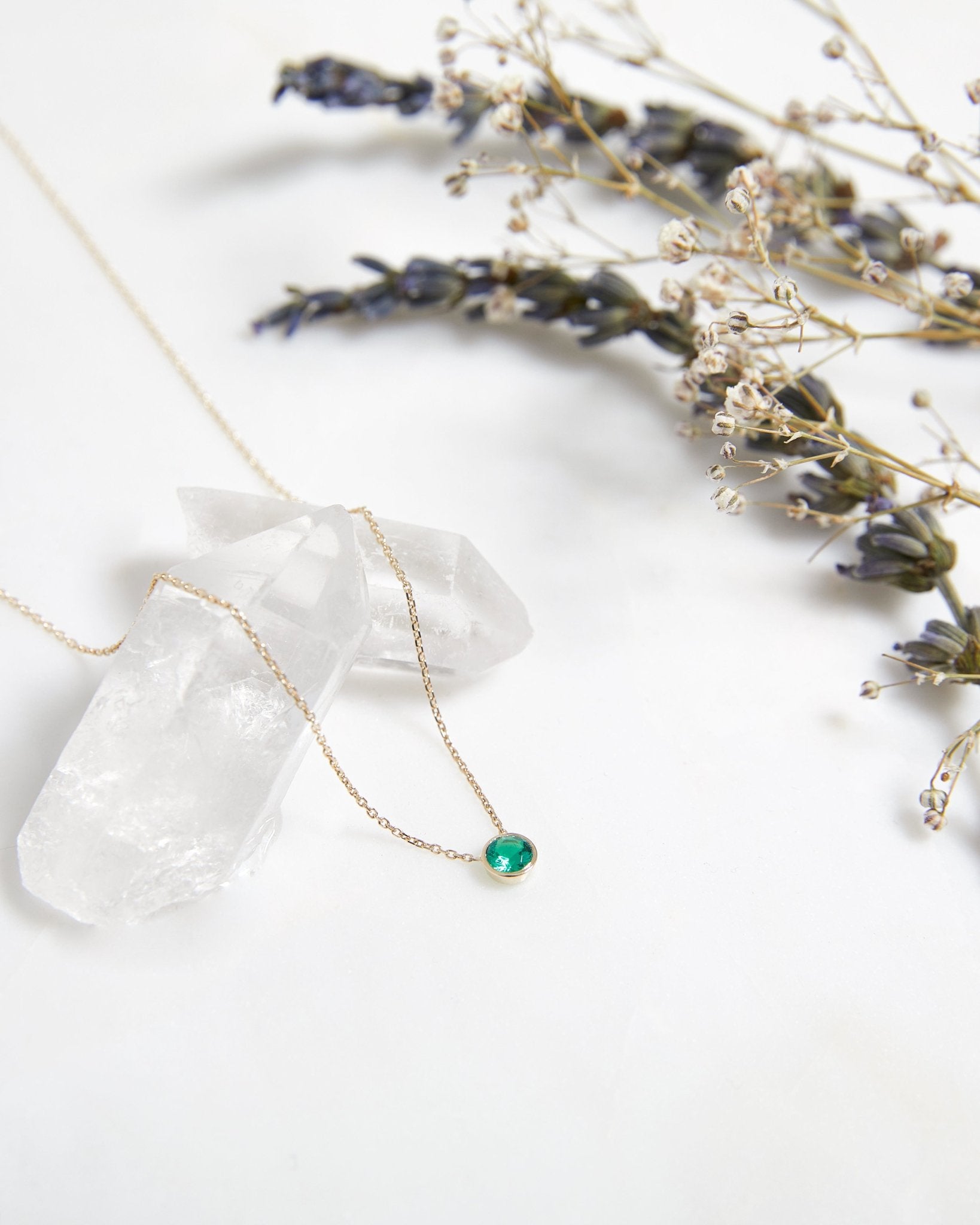 14K solid white gold bezel set solitaire emerald necklace on display with crystals and dried flowers.