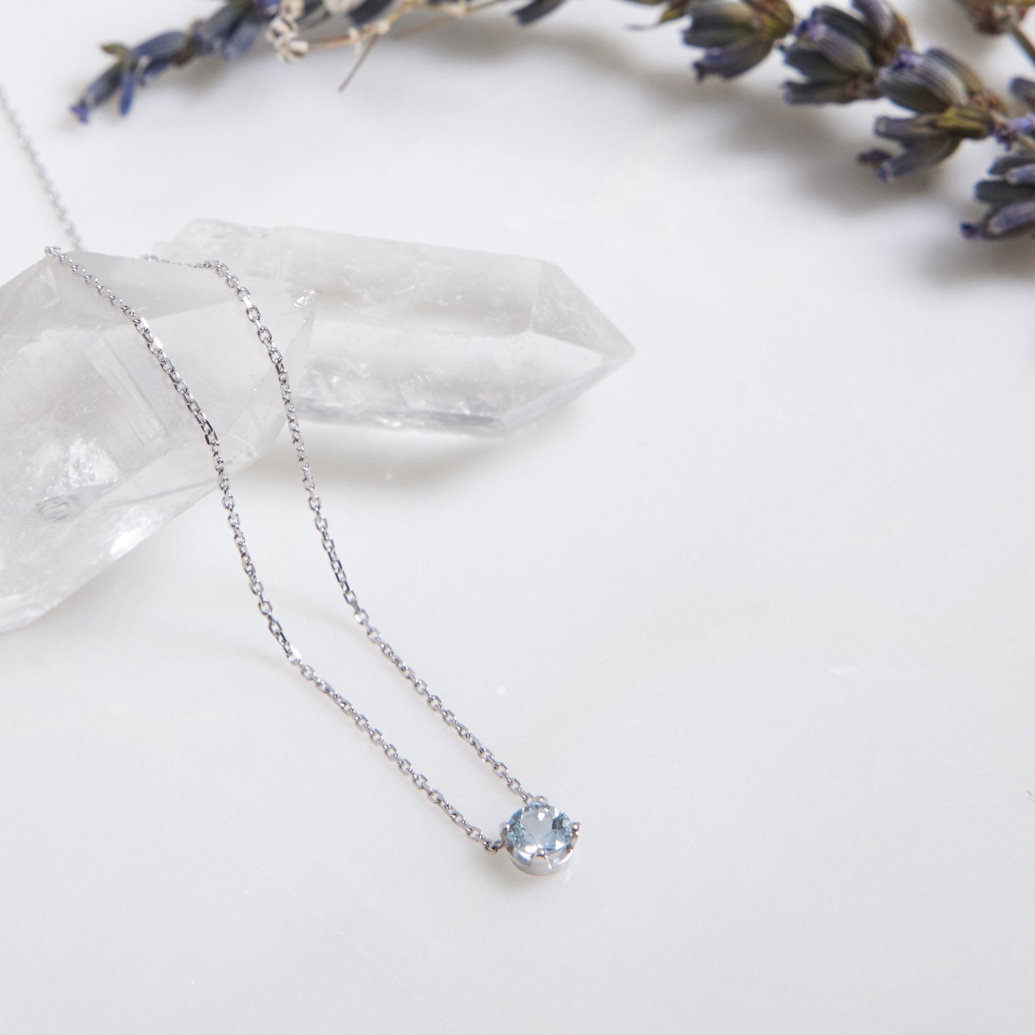 14K yellow gold 5mm prong set solitaire aquamarine necklace on a marble surface with lavender accents.