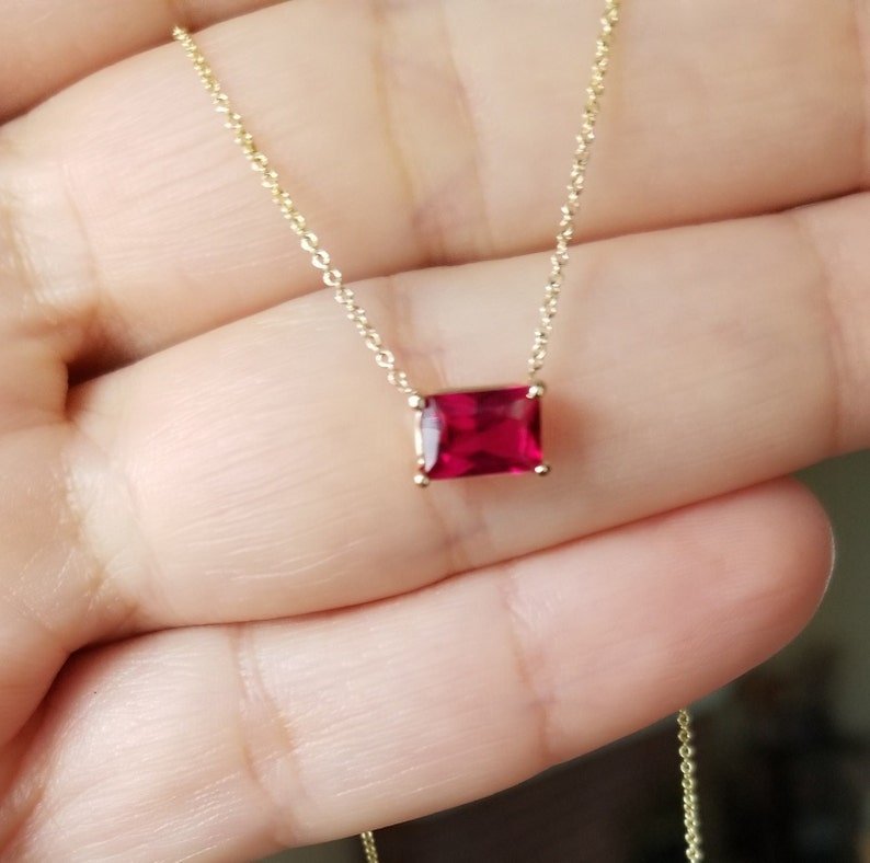 14K white gold emerald cut ruby solitaire necklace displayed on a hand, showcasing the 7x5mm pendant.