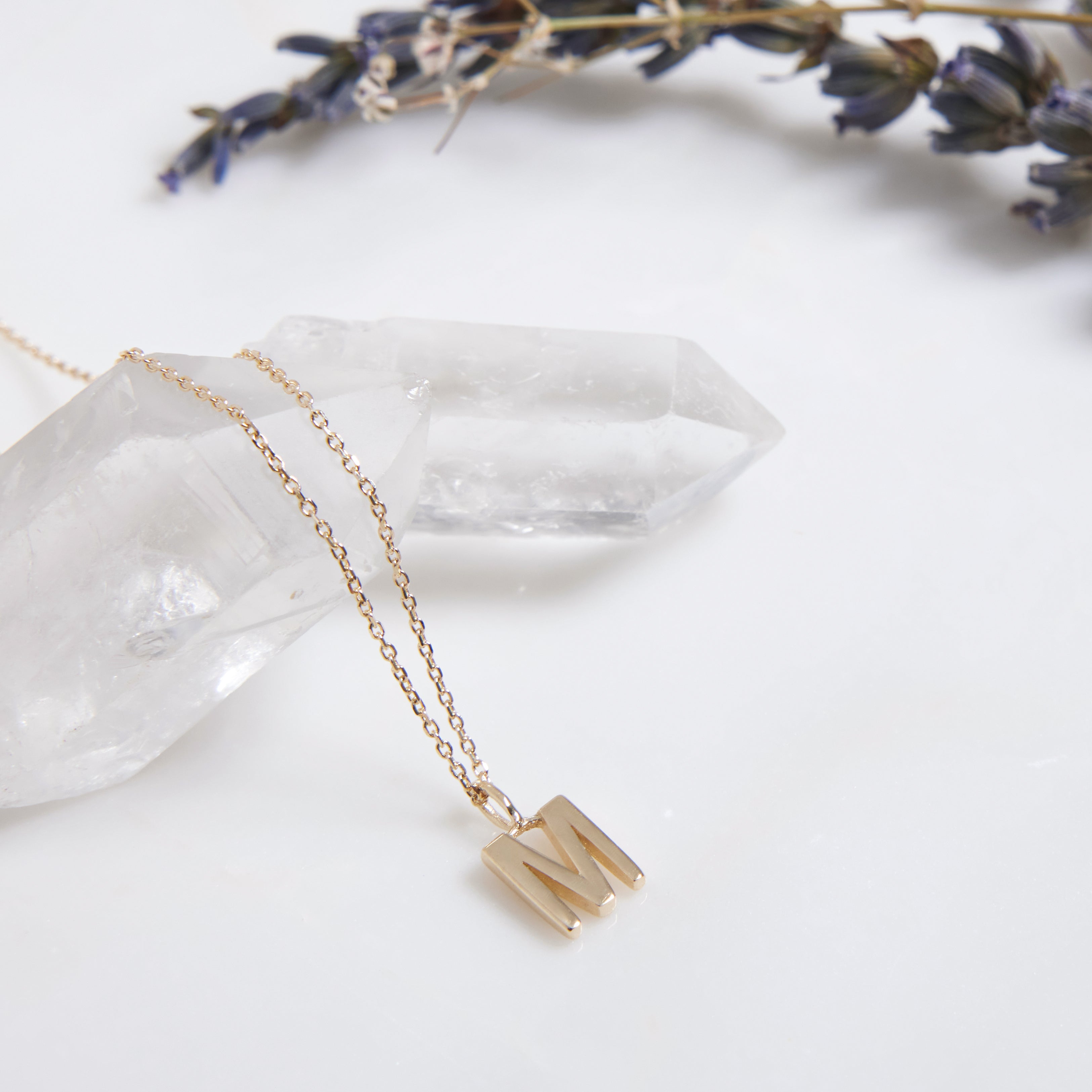 Dainty 14K solid yellow gold initial necklace featuring letter M, displayed on a crystal with lavender sprigs.