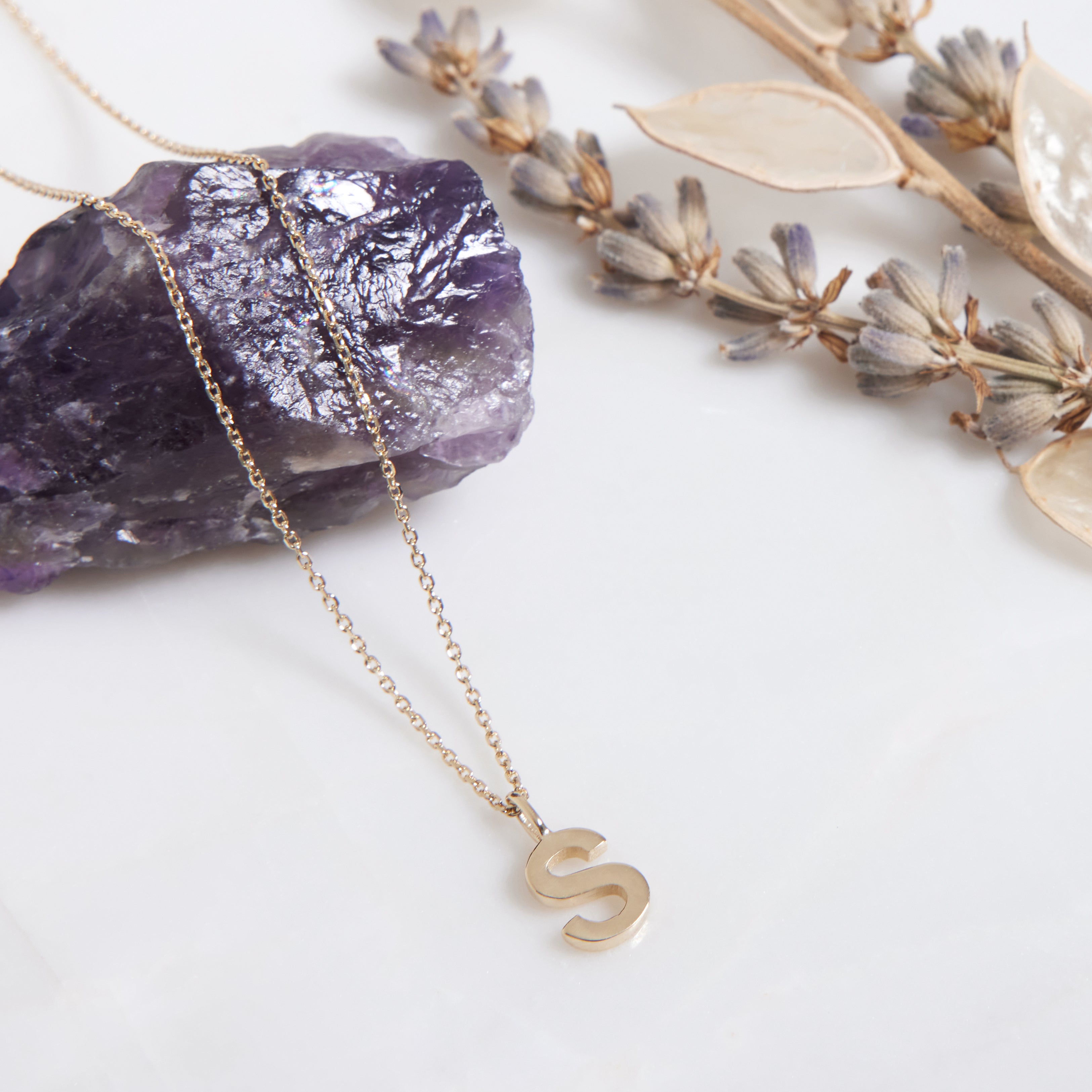 14K solid yellow gold initial necklace with letter S, displayed with a purple crystal and dried flowers.