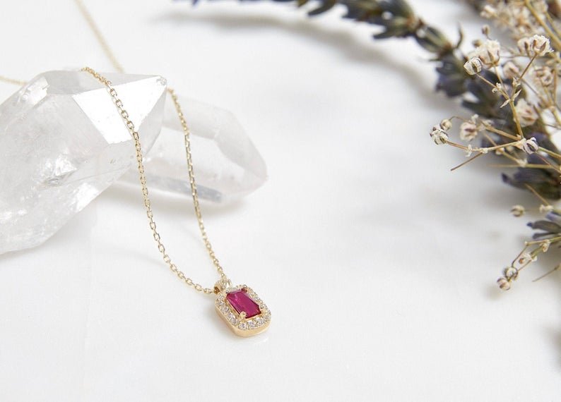 14K yellow gold emerald cut solitaire ruby necklace displayed on a crystal with dried flowers, elegant and dainty design.