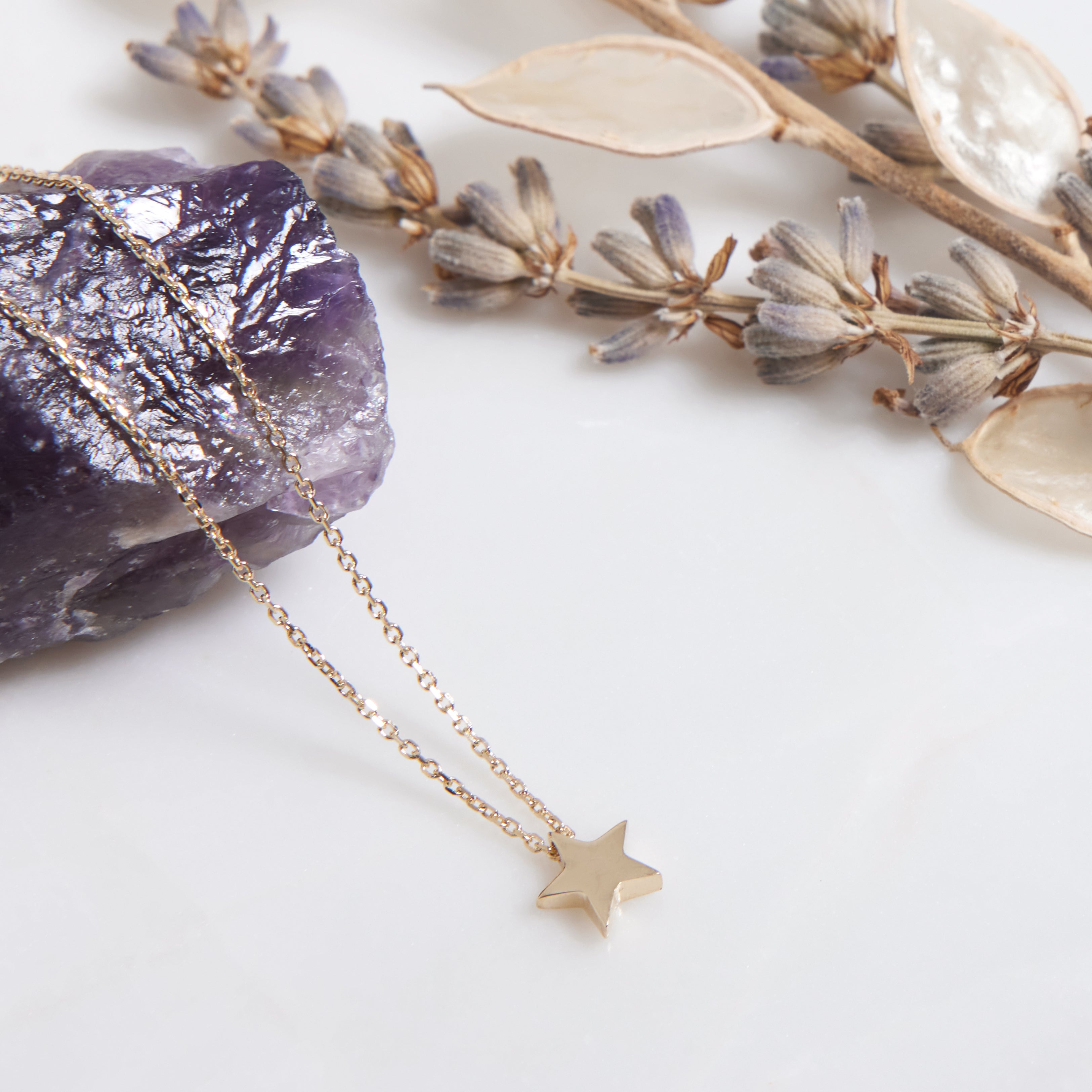 14K solid yellow gold minimalist star necklace displayed on a marble surface with dried flowers and a purple stone.