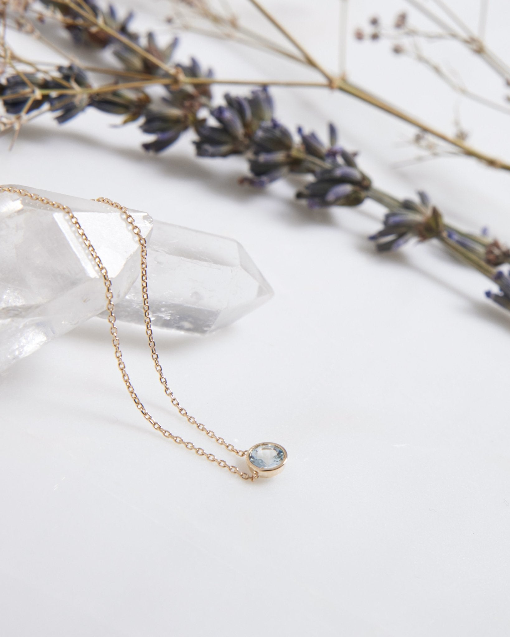14K white gold 6mm bezel set aquamarine solitaire necklace resting on a crystal, with lavender flowers in the background.