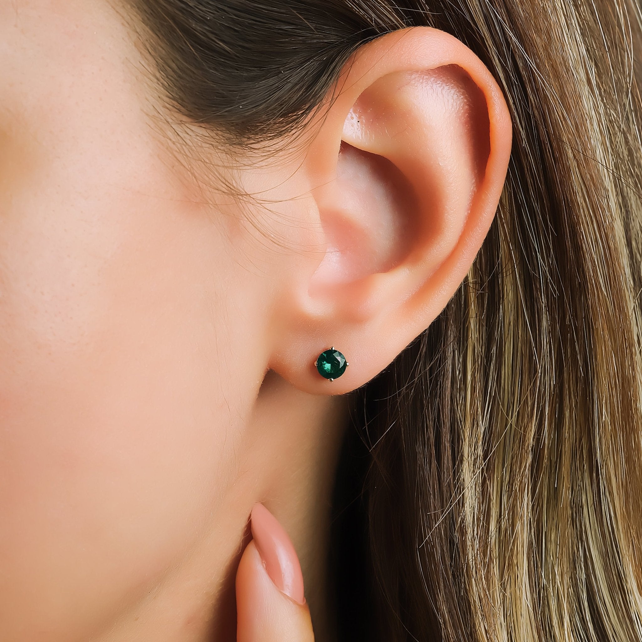 Close-up of a woman's ear wearing 14K yellow gold 5mm round emerald stud earrings, showcasing their elegance.