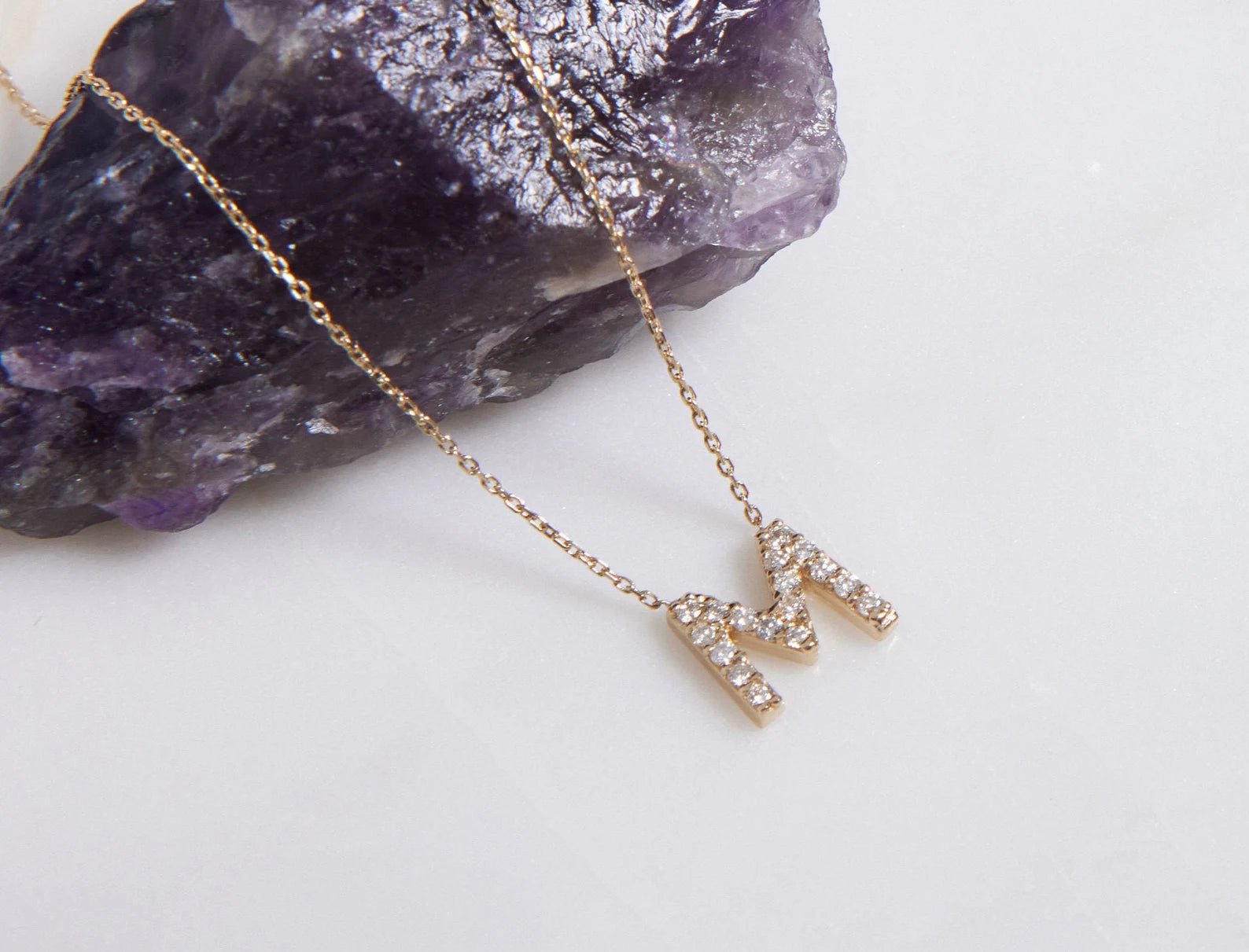 14K yellow gold diamond initial necklace with letter M, featuring sparkling white diamonds, on an amethyst backdrop.