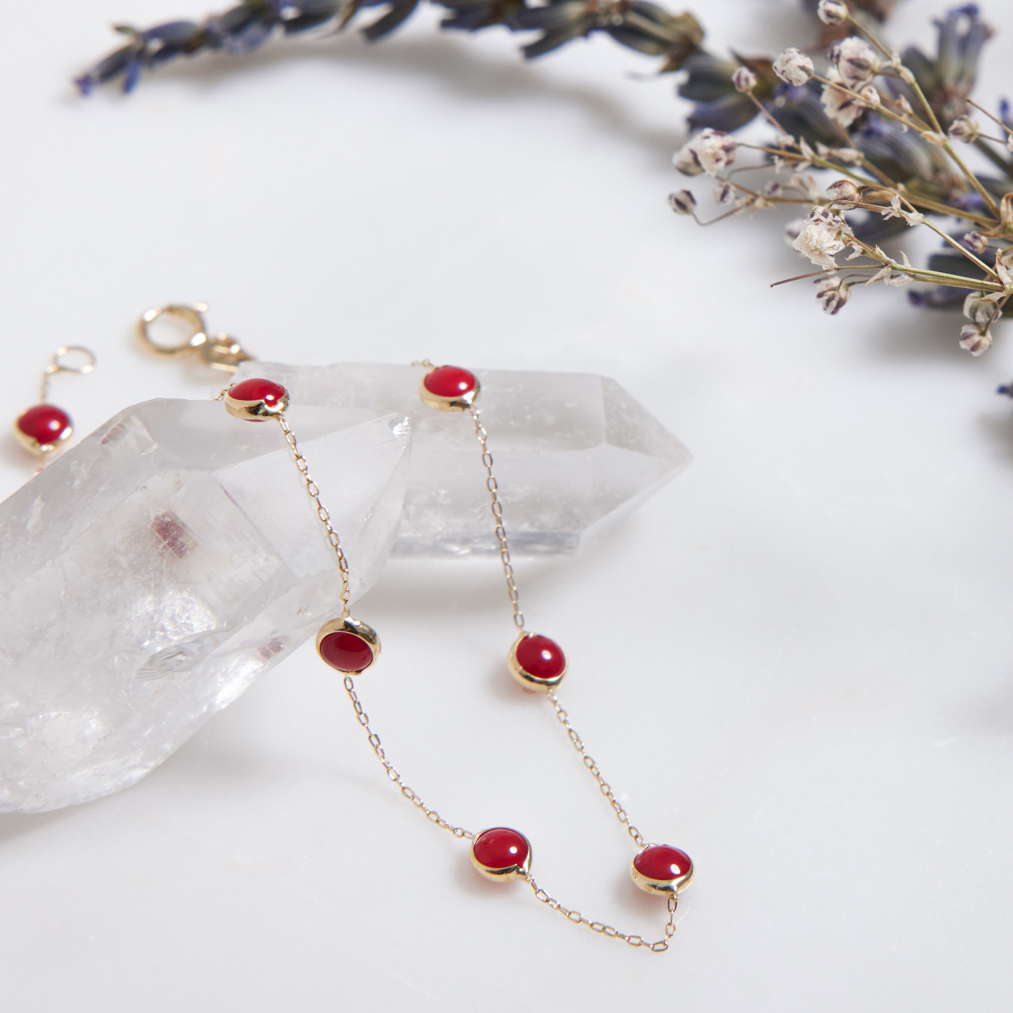 Delicate gold bracelet with red gemstones, resting on clear quartz crystal and lavender sprigs, showcasing elegant jewelry design.