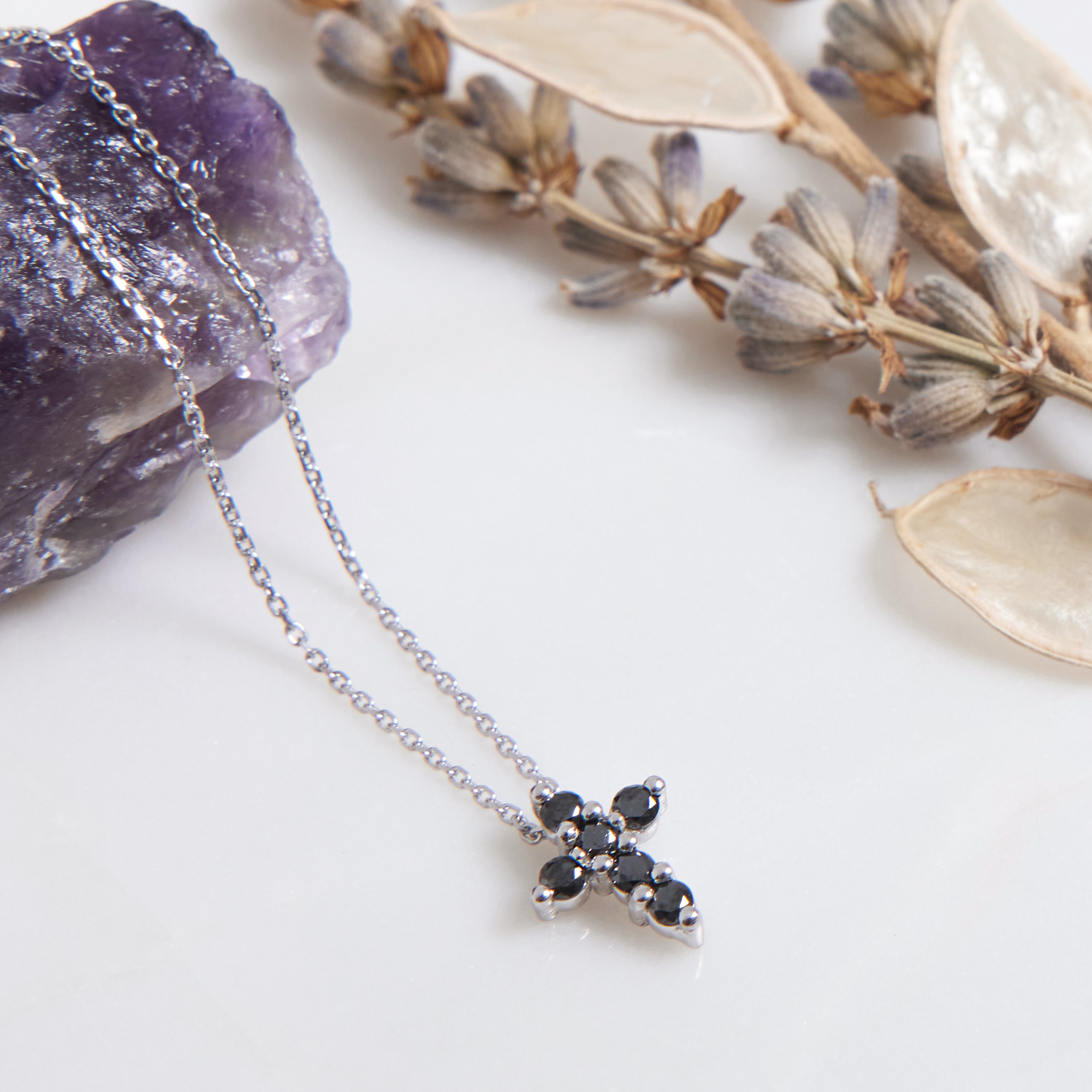 14K solid white gold cross necklace with black diamonds, placed on a marble surface with amethyst and dried flowers.