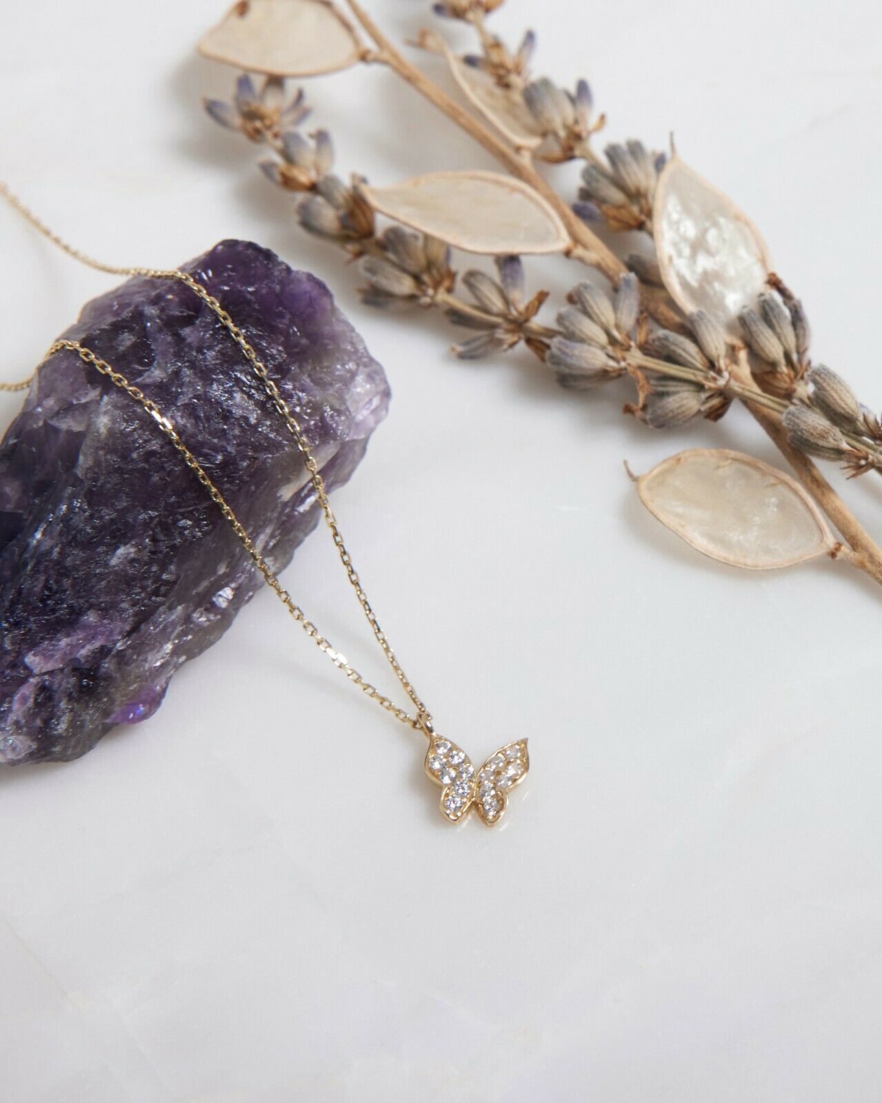14K solid yellow gold diamond CZ butterfly necklace resting on amethyst stone with dried flowers.