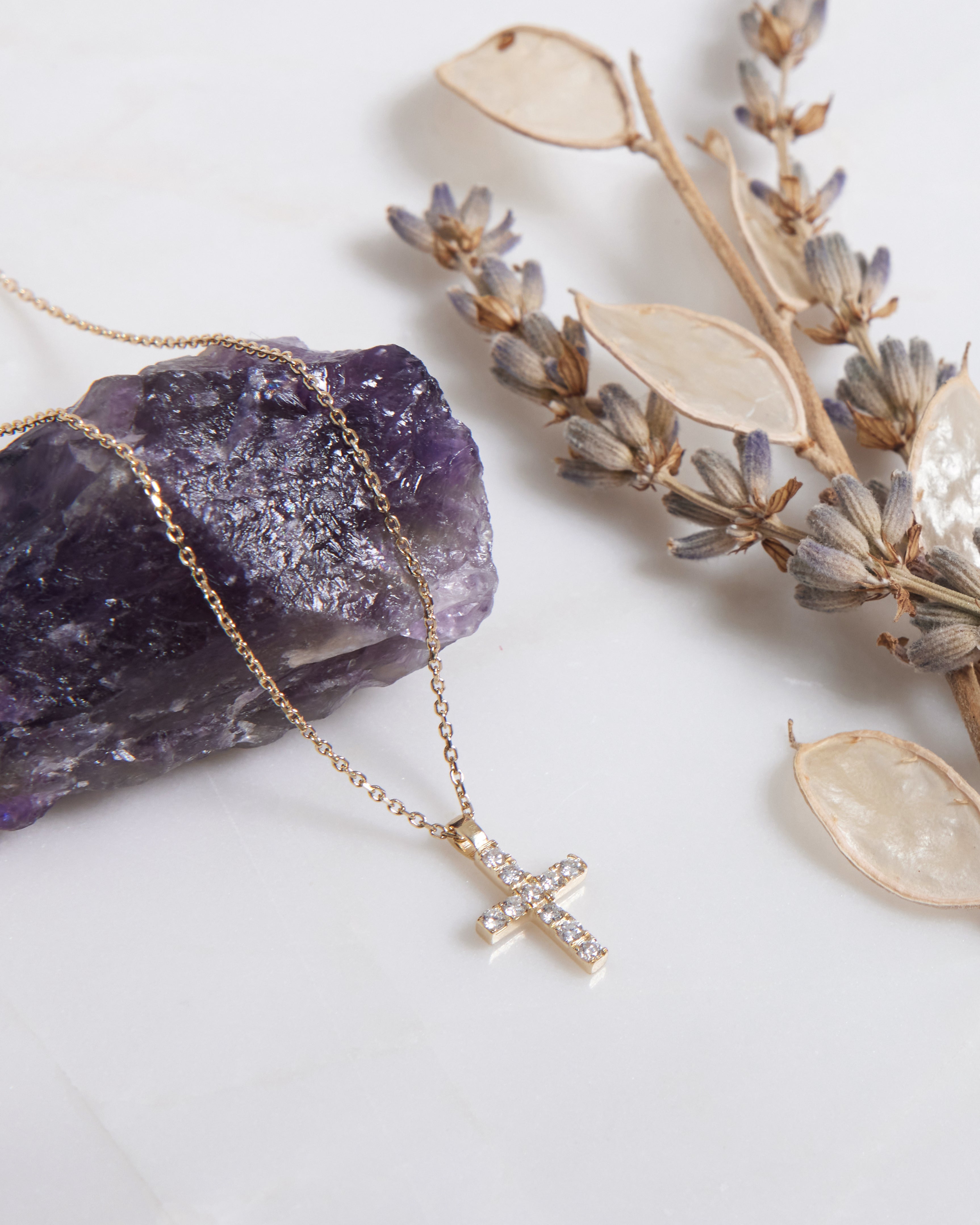 14K yellow gold minimalist diamond cross necklace displayed with purple crystal and dried flowers.