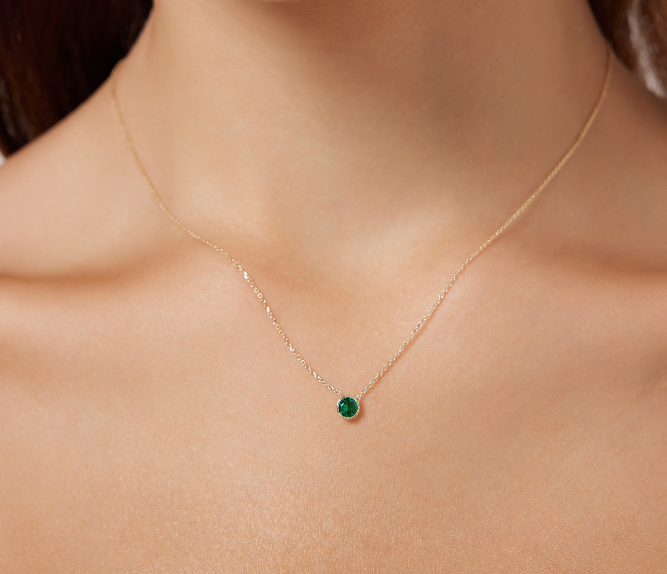 14K solid yellow gold 4mm bezel set emerald solitaire necklace displayed on a model's neck.