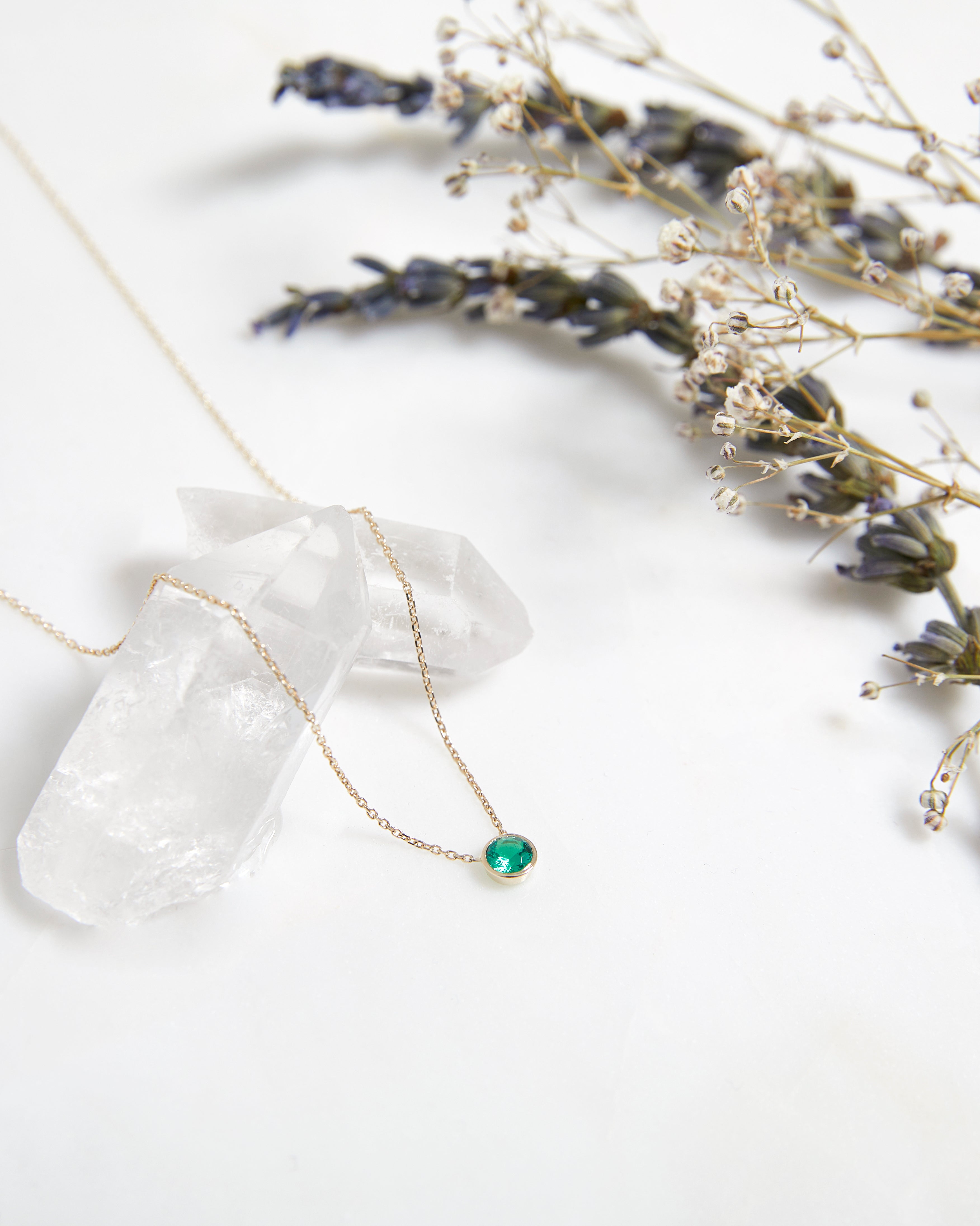 14K solid yellow gold minimalist emerald solitaire necklace on crystal with dried flowers backdrop.