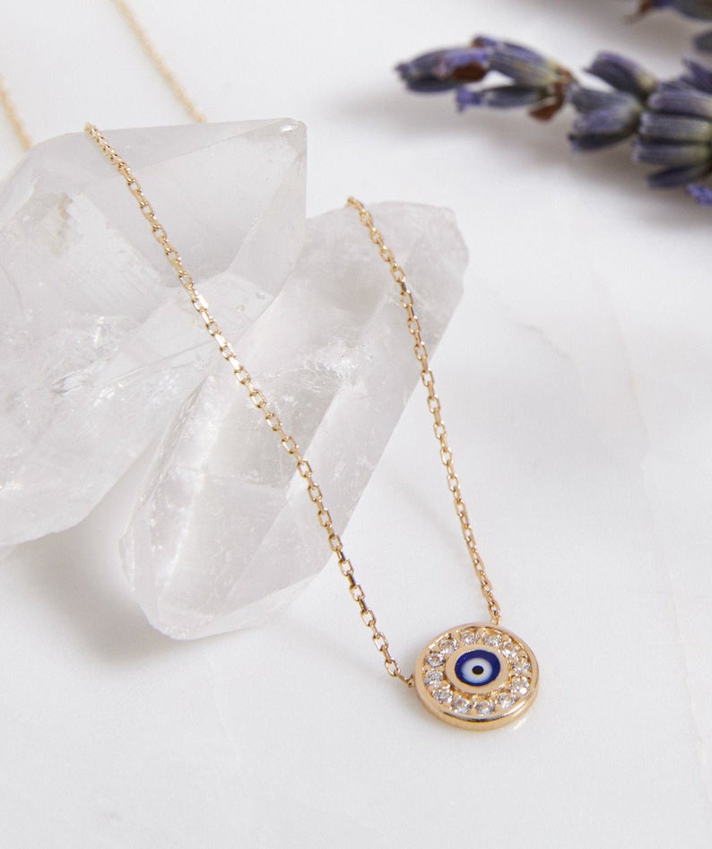 14K solid yellow gold minimalist round evil eye necklace with CZ, resting on clear quartz crystal.