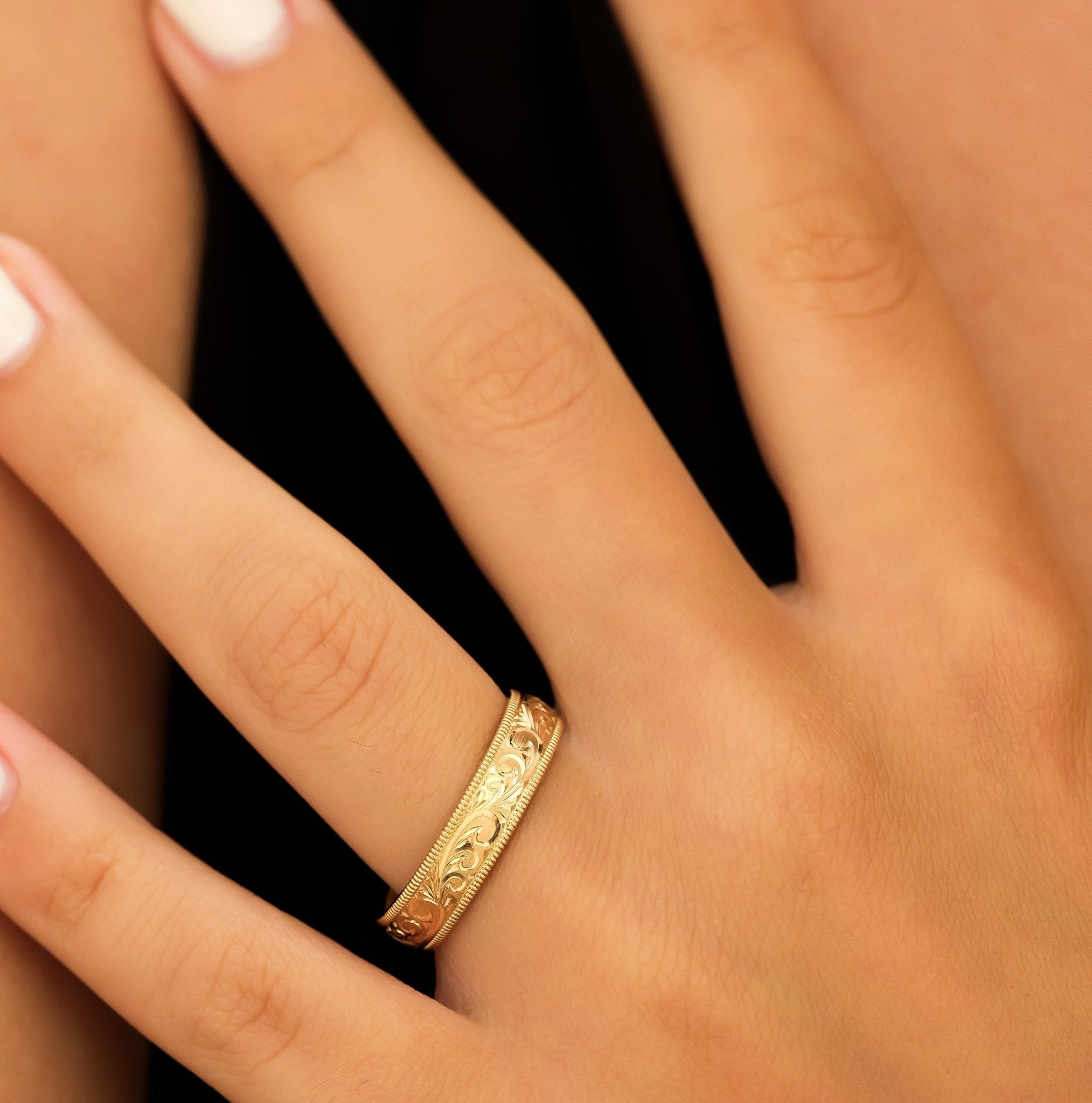 Hand engraved yellow gold men's wedding band displayed on a finger, featuring intricate leaf motifs and shiny finish.