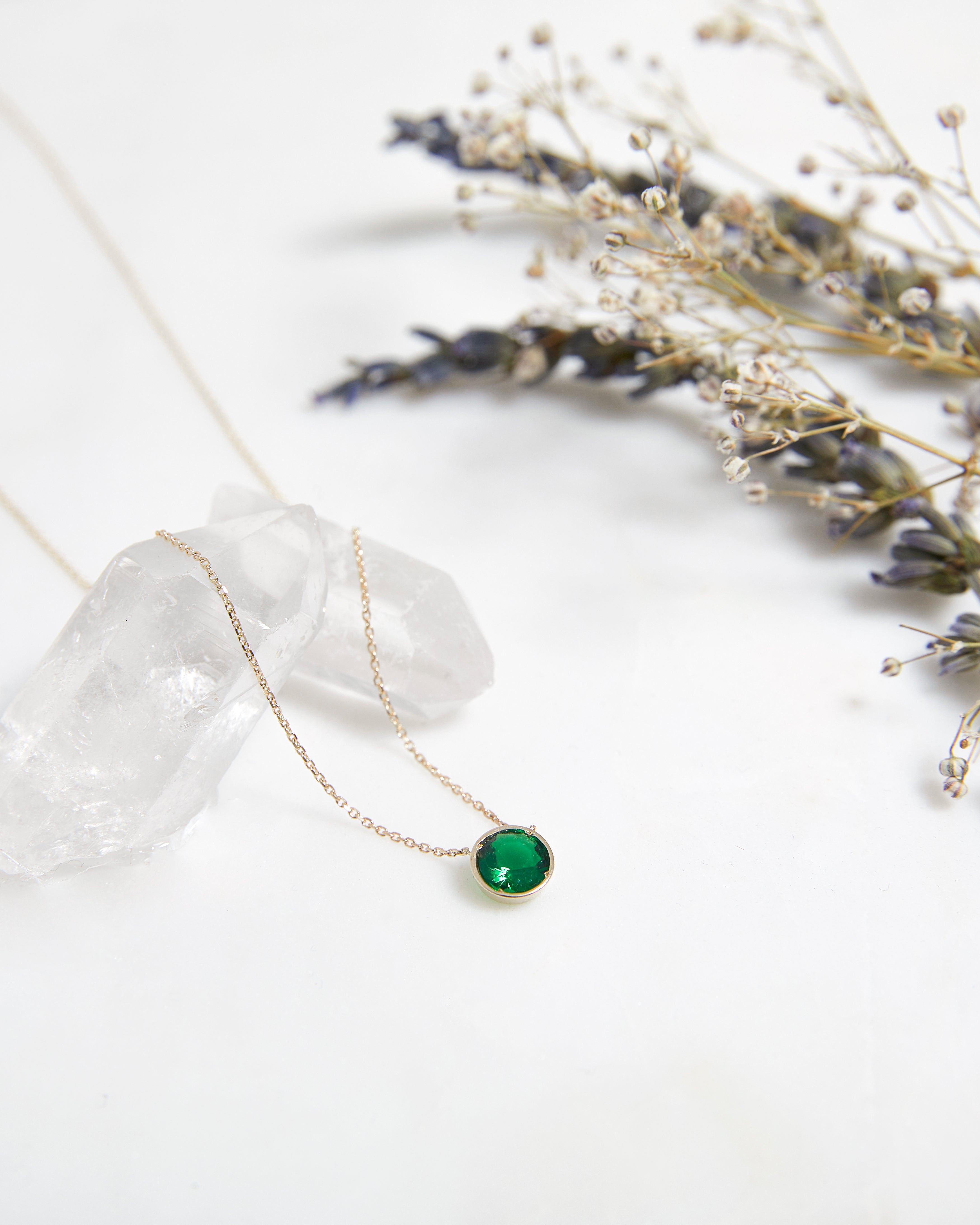 14K yellow gold 6mm bezel set emerald solitaire necklace with a delicate chain, styled with dried flowers.