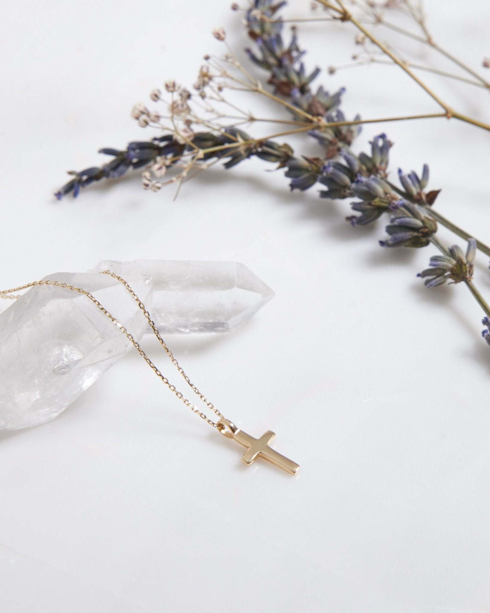 14K solid yellow gold cross necklace resting on a crystal, surrounded by delicate flowers on a marble surface.
