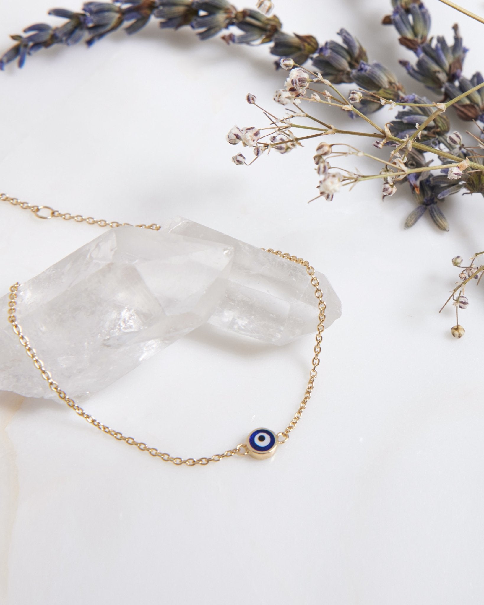 14K solid yellow gold minimalist evil eye bracelet with a blue and white charm, placed on a crystal and floral backdrop.