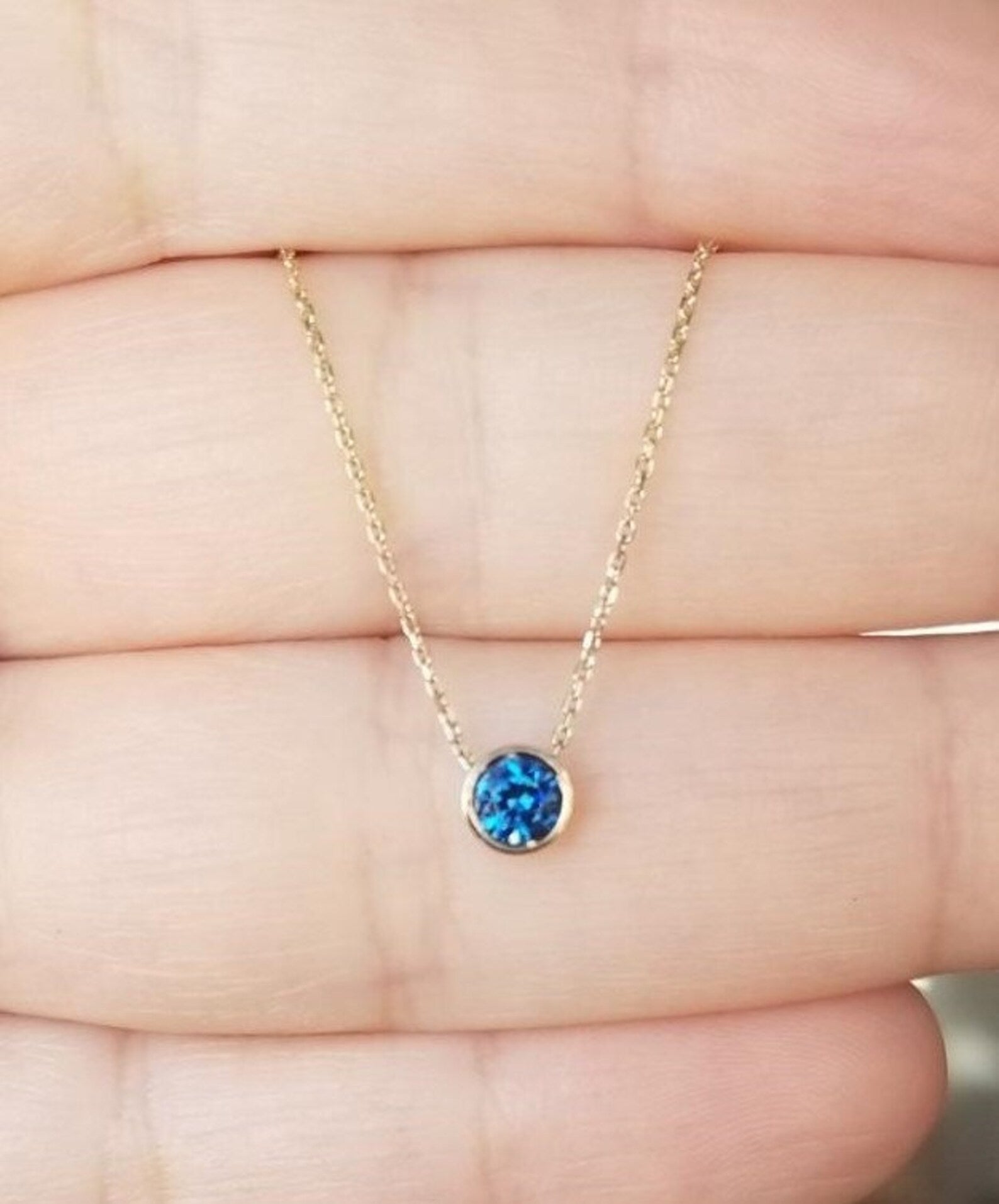 14K yellow gold 4mm bezel set sapphire necklace displayed on a hand, showcasing its minimalist design and vibrant blue stone.