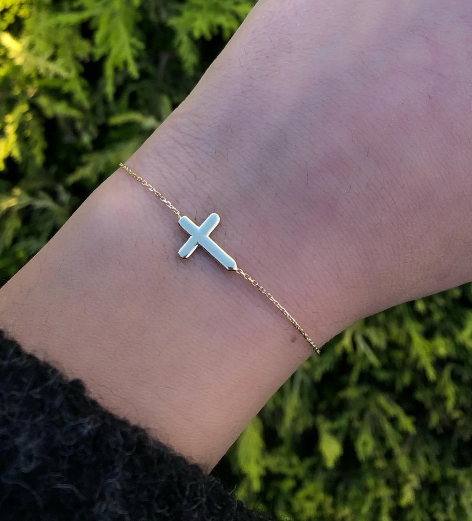 14K solid yellow gold sideway cross bracelet worn on wrist surrounded by greenery, showcasing minimalist design.