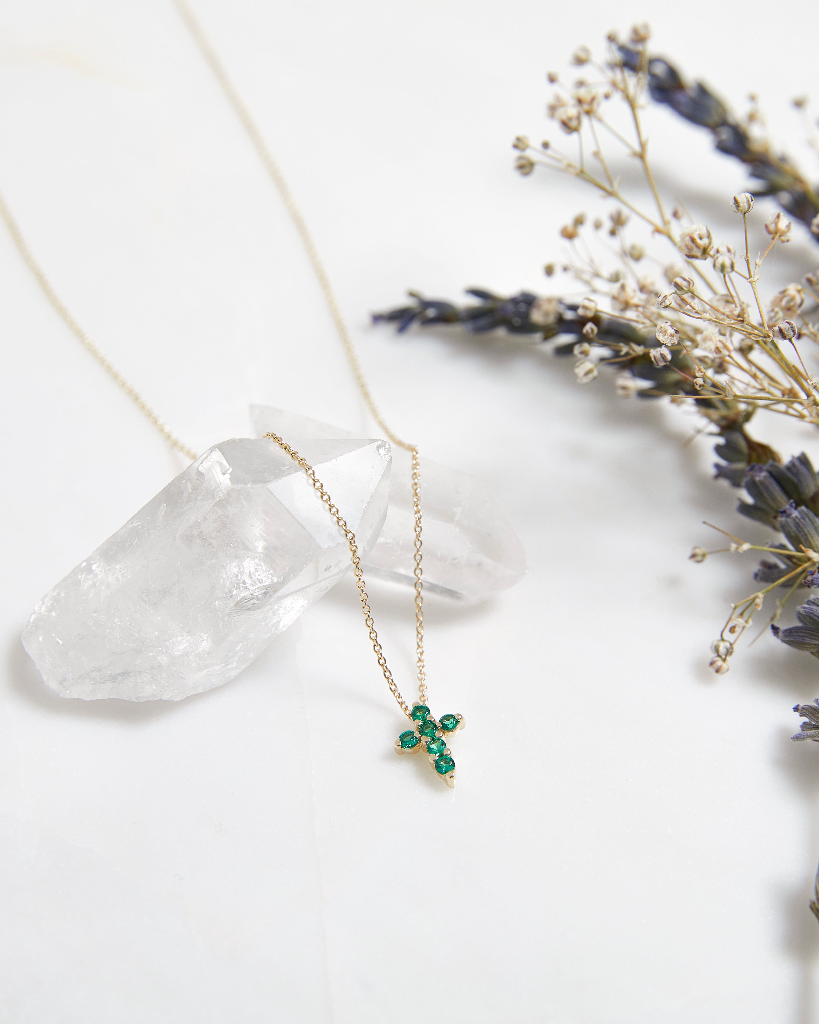 14K solid yellow gold minimalist cross necklace with emeralds, placed on a crystal with dried flowers.