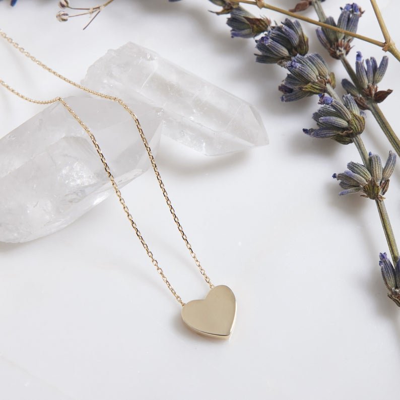 14K yellow gold minimalist heart necklace on a marble surface with lavender and crystals.