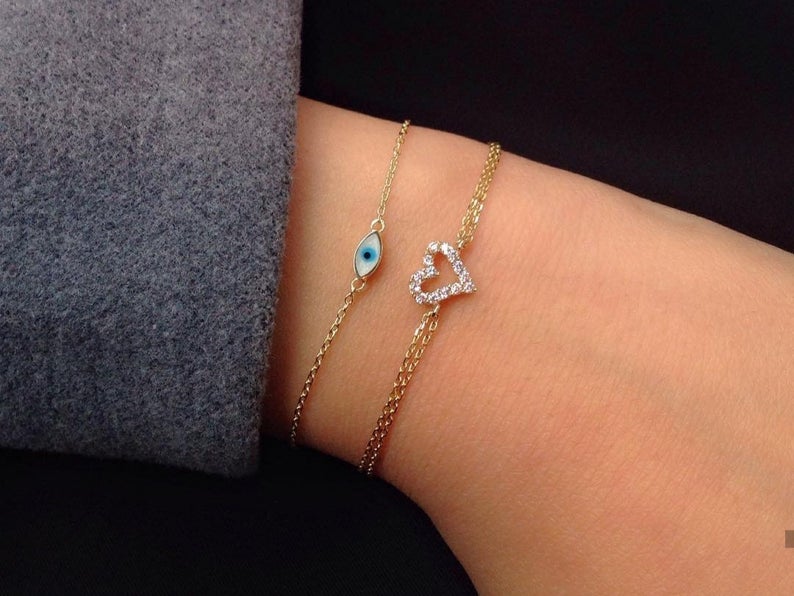 14K yellow gold minimalist evil eye bracelet with blue enamel, paired with a dainty heart bracelet on a wrist.