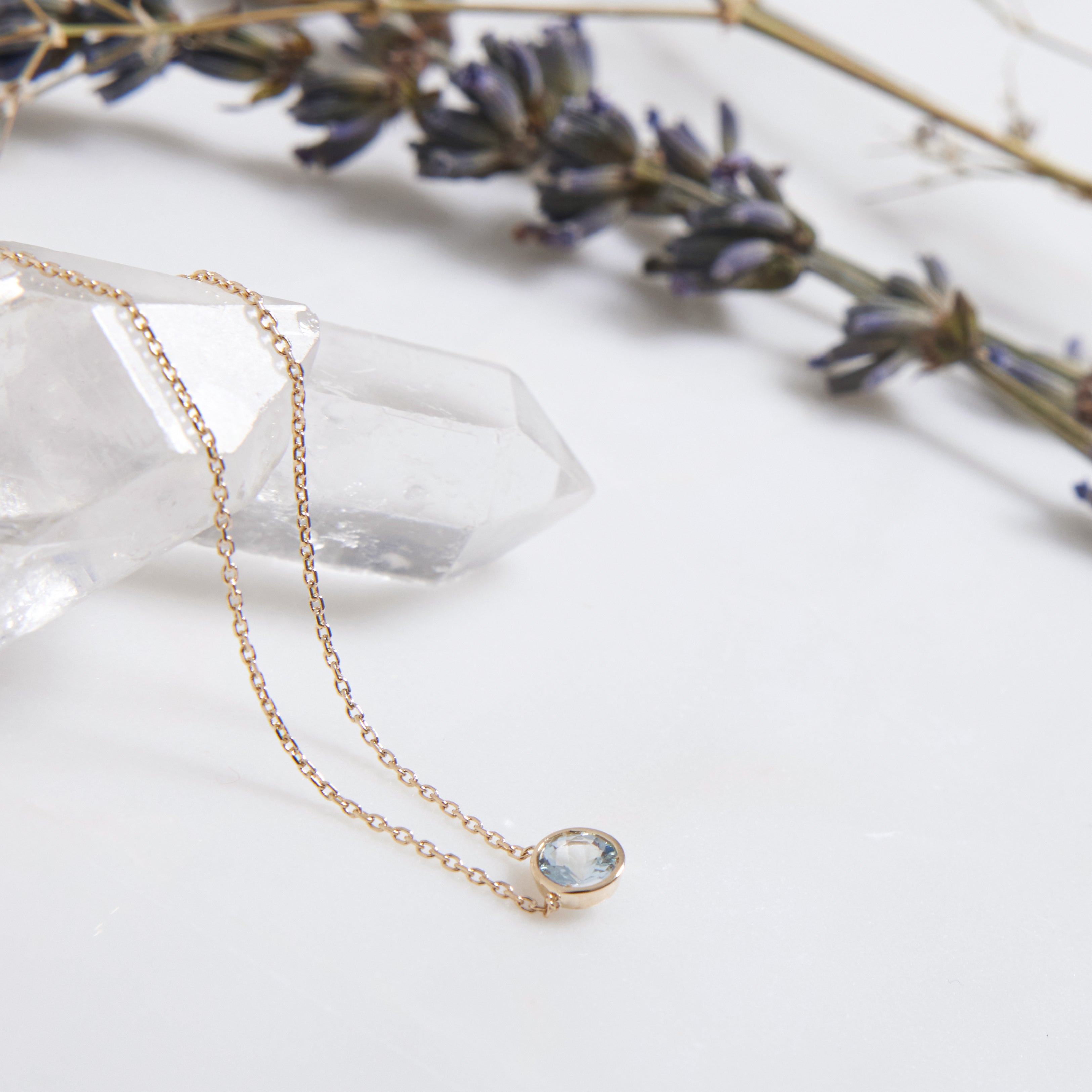 14K yellow gold minimalist aquamarine solitaire necklace with 4mm bezel setting, placed on a crystal with lavender.