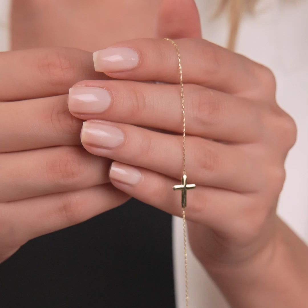 Dainty 14K solid yellow gold sideway cross bracelet held delicately between fingers, showcasing minimalist elegance.