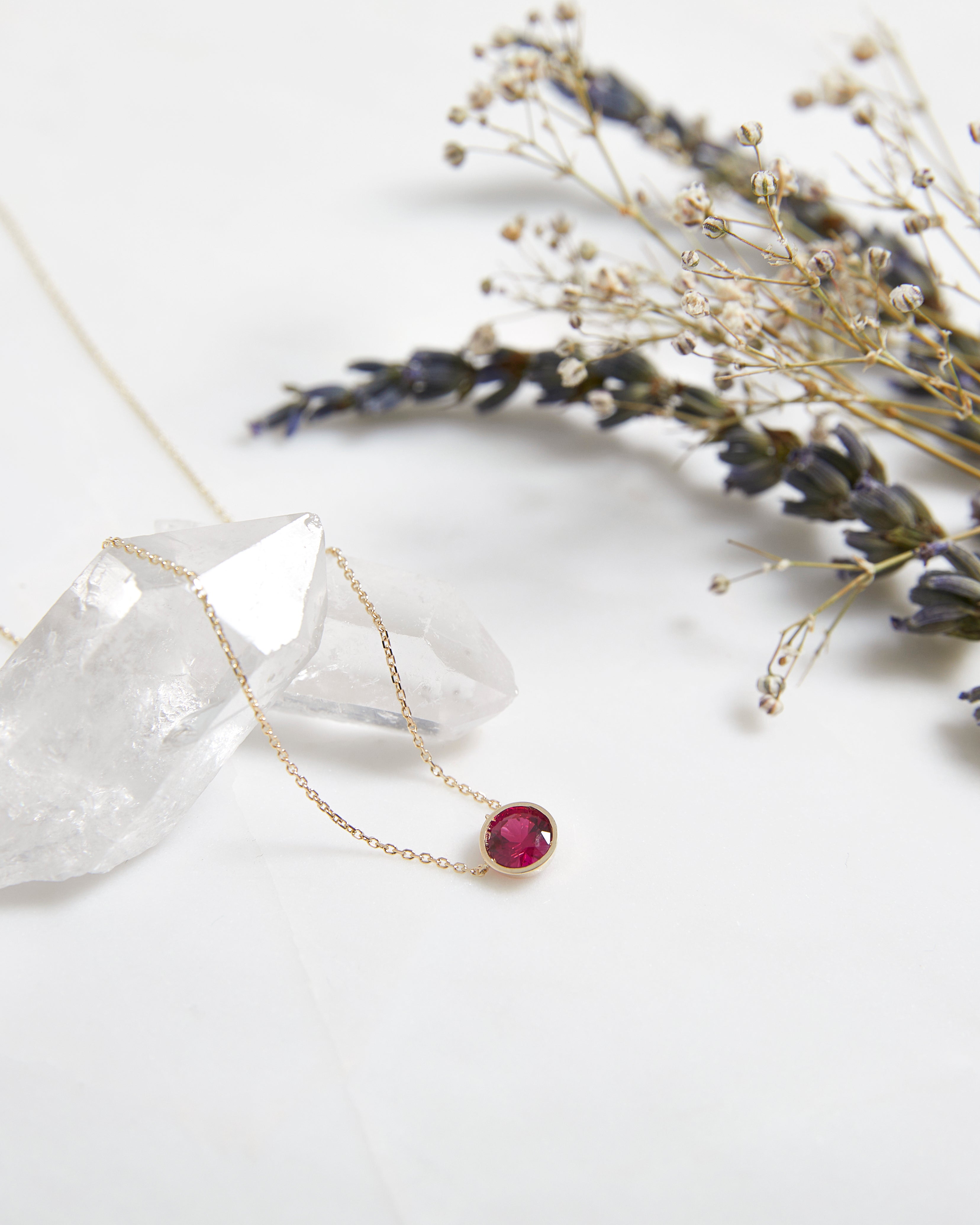 14K yellow gold bezel set ruby solitaire necklace on crystal with dried flowers, perfect for everyday elegance.